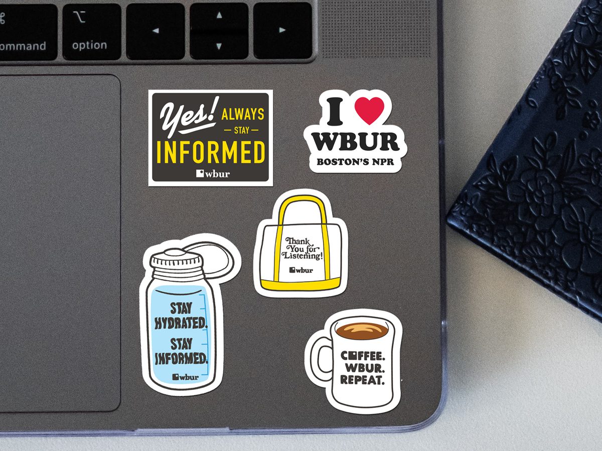 A laptop decorated with five WBUR-themed stickers, including phrases like “Stay Hydrated. Stay Informed.”, “Yes! Always Stay Informed”, and “Coffee. WBUR. Repeat.” The stickers are arranged near the keyboard and trackpad.